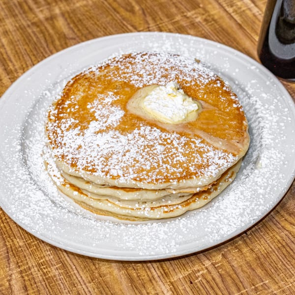 Stack of golden pancakes with melting butter, syrup, and powdered sugar on a white plate.