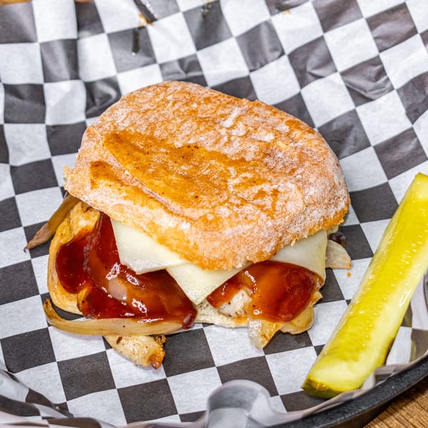 BBQ chicken sandwich with melted cheese and onions, served with a pickle spear on checkered paper.