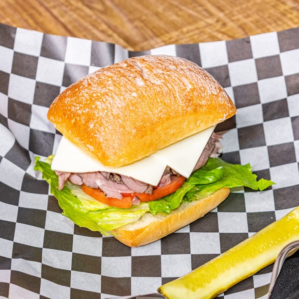 Roast beef sandwich with cheese, lettuce, and tomato on a ciabatta bun, with a pickle spear.