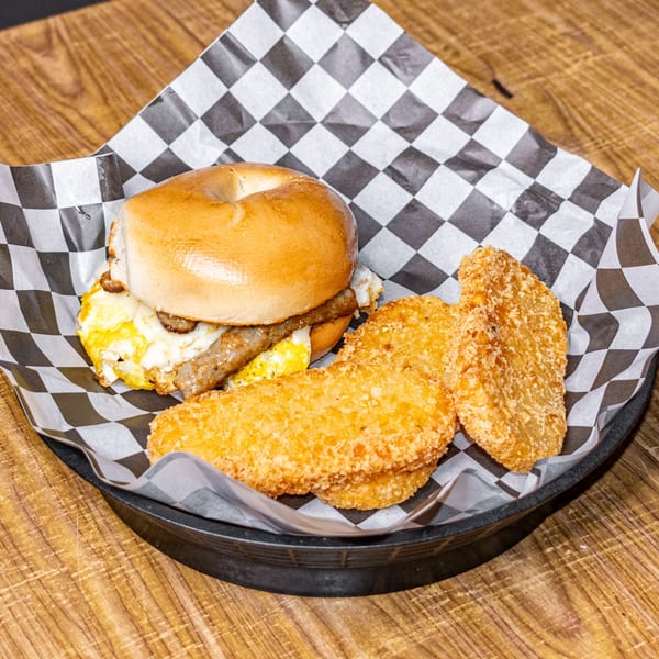 Breakfast bagel sandwich with sausage and egg, served with three hash browns in a basket.