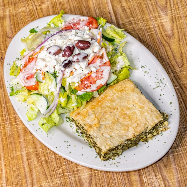 A white plate with a slice of spanakopita and a Greek salad topped with feta, olives, and dressing.