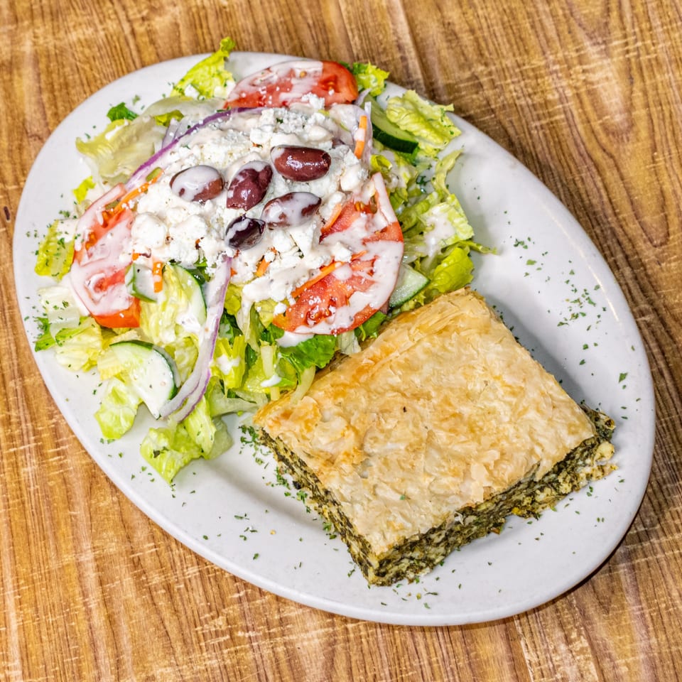 A white plate with a slice of spanakopita and a Greek salad topped with feta, olives, and dressing.
