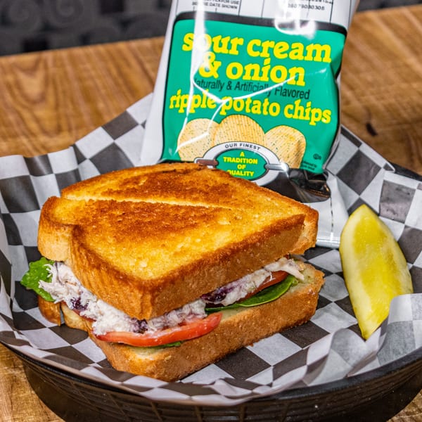 Grilled sandwich with chicken salad, tomato, lettuce, a pickle spear, and sour cream & onion chips.